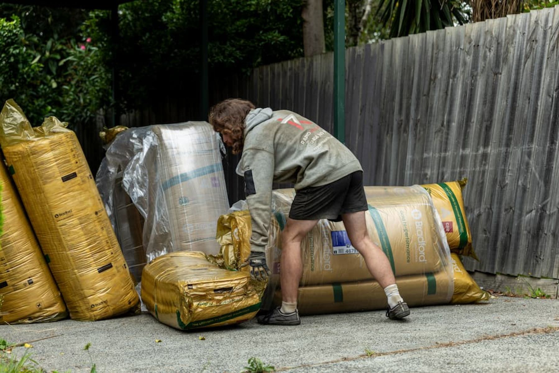 Worker handling ceiling insulation batts stacked on a residential driveway prior to installation.