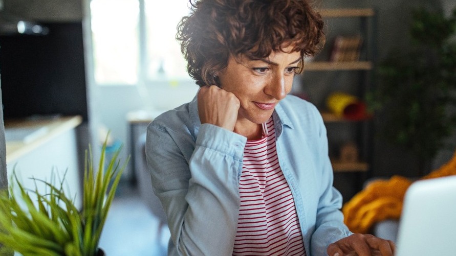 A woman with brown hair sits in front of a laptop. A couch and a bookshelf are behind her. 