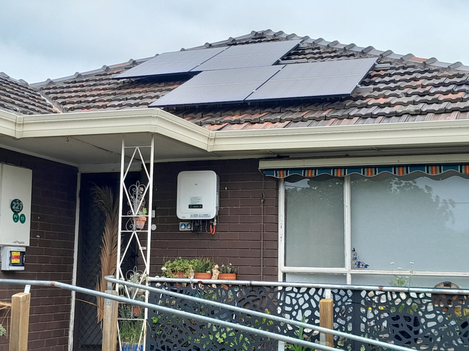 Residential house with solar panels installed on a tiled roof, showcasing renewable energy use. The house features a brown brick exterior, a white electrical box mounted on the wall, potted plants on a ledge, and a metal railing in front.