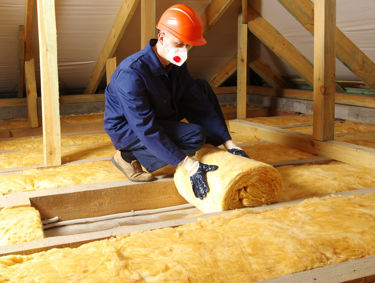 Person wearing safety gear installing yellow fibreglass insulation between timber attic joists