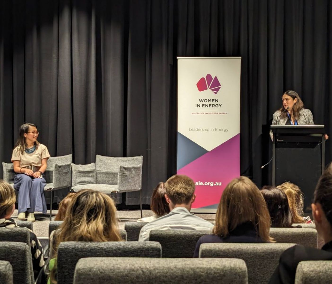 A person presents at a women in energy conference while another is seated on stage