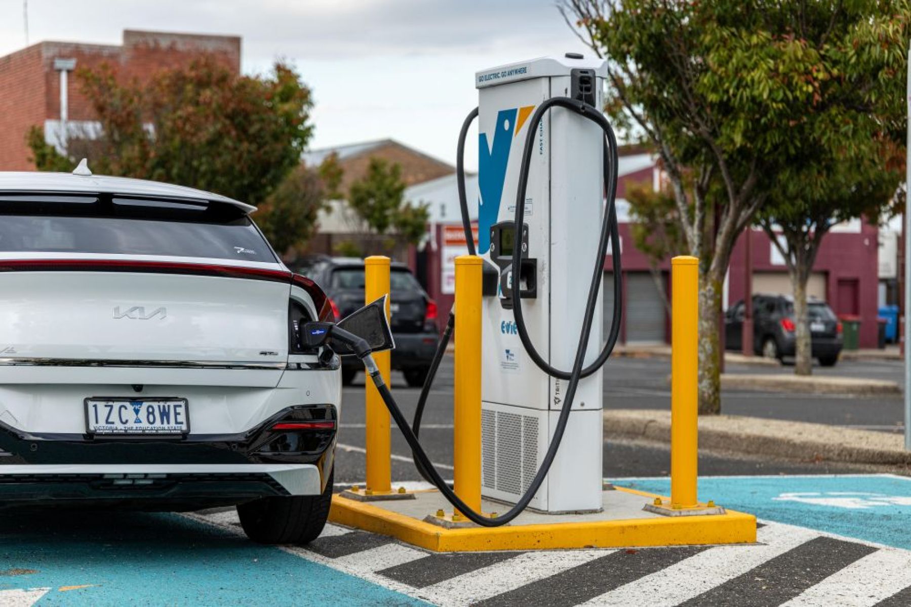 Photo of the back of a white car connected to a public electric vehicle charger in a carpark with trees and other cars in the background. 