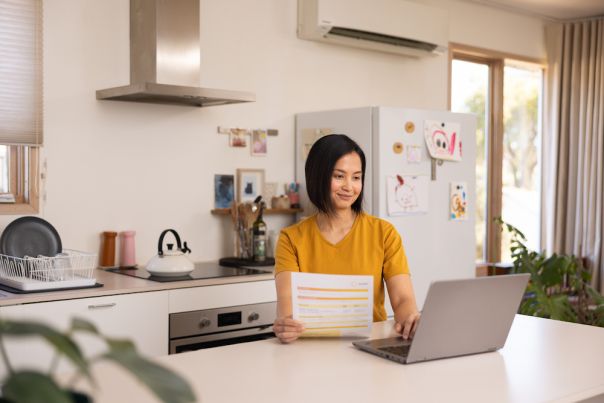 Woman sitting at kitchen bench looking at laptop and paper energy bill