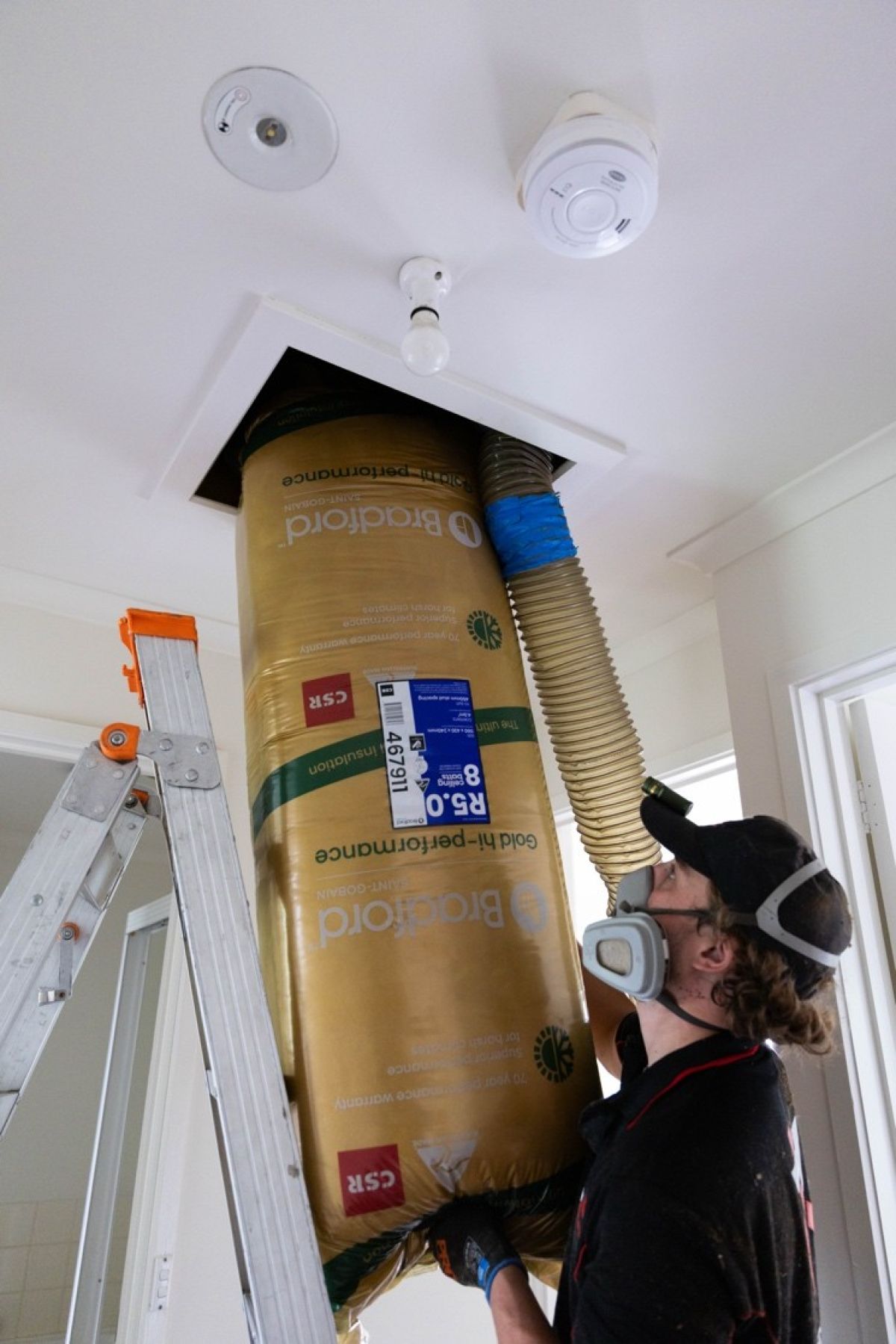 Worker wearing protective goggles and respirator stands beside an aluminum ladder loading insulation into a roof cavity