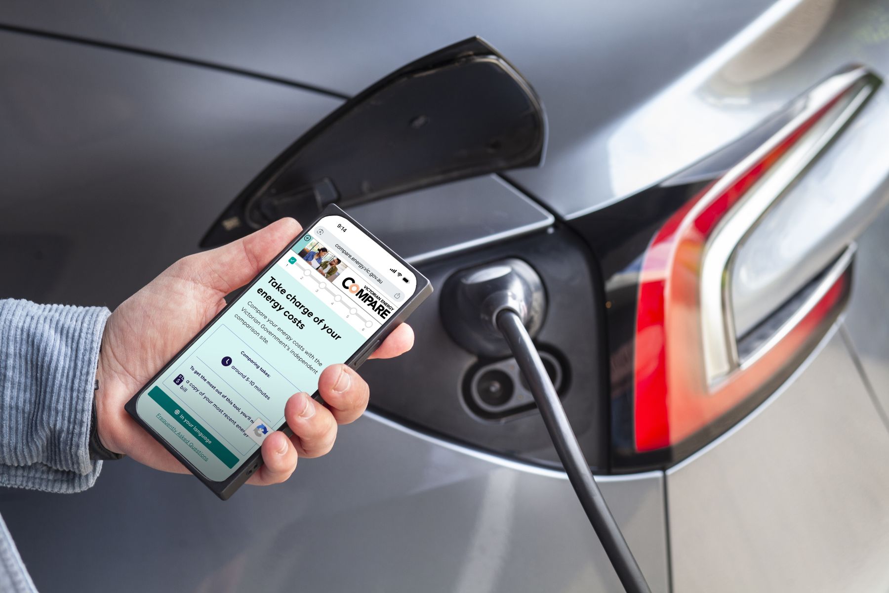 Photo of someone holding a mobile phone with an electric vehicle in the background.