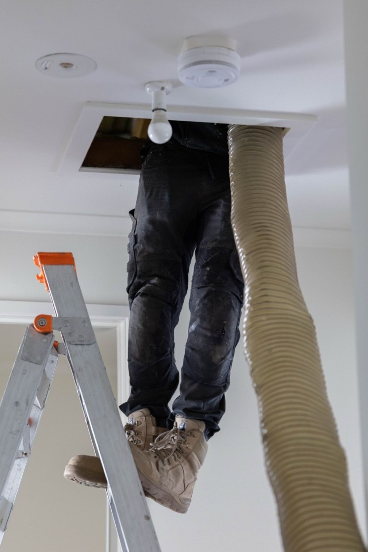 Image showing a worker’s legs extending through a ceiling opening beside a large flexible ventilation duct and step ladder