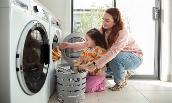 Parent and child sitting on floor loading clothes into a washing machine